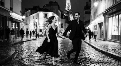 A romantic, grainy black and white photograph of a couple running through a Parisian street at night. The city lights are blurred in the background, creating a soft bokeh effect. The couple's joy is palpable as they hold hands, their attire caught in a motion-blur that gives the image a timeless, cinematic quality. The overall mood is one of pure, unrestrained romance and high-fashion elegance.