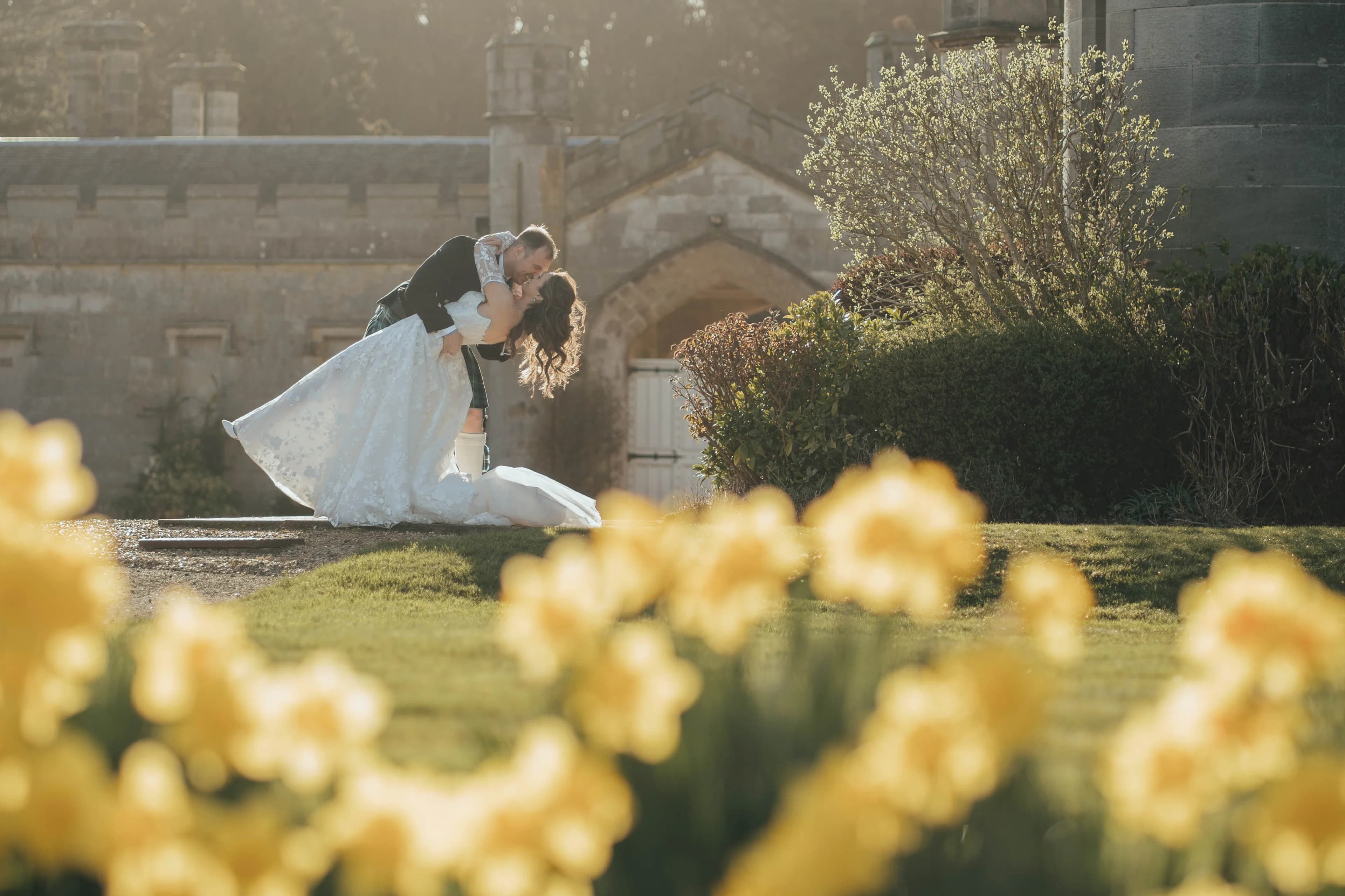 Backlit portrait of a bride a groom kissing in the hazy evening light. There are yellow daffodils in the foreground, and a warm hazy glow over everything. 