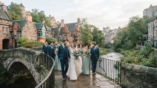 A wide-angle, cinematic photograph of a wedding party walking across a bridge in Edinburgh's Dean Village. The colorful historic buildings and the flowing Water of Leith provide a picturesque backdrop. The image is captured in a documentary style, with natural movements and candid expressions, using a soft, cool-toned color palette that evokes a serene Scottish morning.