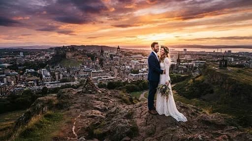 A breathtaking full-width landscape photograph of a couple standing on the summit of Arthur's Seat at sunset. The entire city of Edinburgh is spread out below them, bathed in the golden, orange, and purple hues of the setting sun. The composition is expansive and epic, capturing the rugged natural beauty of the Scottish landscape combined with the urban silhouette of the historic city.
