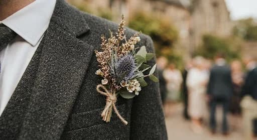 A minimalist, close-up photograph of a groom's boutonniere featuring local Scottish thistles and dried heather, pinned to a textured charcoal tweed suit. The image is focused and crisp, with a shallow depth of field that blurs the background. The aesthetic is modern and refined, highlighting the connection to Scottish tradition through high-end photography.