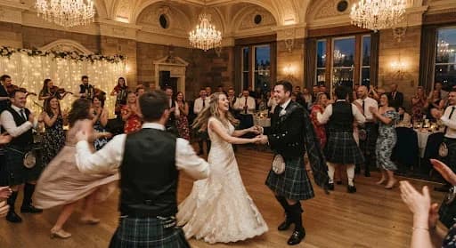 A joyful and energetic photograph of a traditional Scottish Ceilidh dance during a wedding reception in a grand ballroom in Edinburgh. The movement is captured with a slight motion blur to convey the excitement and rhythm of the dance. The warm, ambient lighting of the chandeliers creates a festive and inviting atmosphere, full of laughter and celebration.
