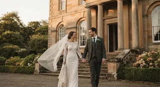 A cinematic, high-end wedding portrait taken at the historic Callendar House in Falkirk. The architecture features grand stone pillars and lush greenery, bathed in soft, golden hour light. The couple is elegantly dressed, with the bride's lace veil catching a gentle breeze. The overall mood is archival, romantic, and exclusive, reflecting a fine-art editorial photography style with soft contrast and warm, neutral tones.