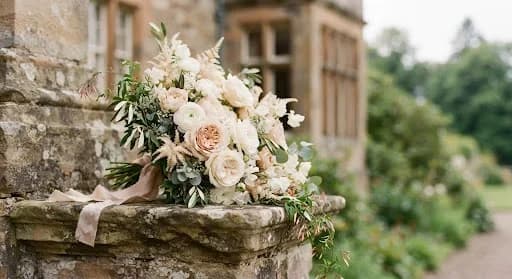 An editorial, close-up photograph of a wedding bouquet resting on an antique stone ledge at Callendar House. The flowers are in shades of cream and blush, with delicate eucalyptus leaves. The focus is sharp on the textures of the petals and the weathered stone, while the background is softly blurred. The lighting is natural and soft, evoking a sense of timeless elegance and quiet luxury.
