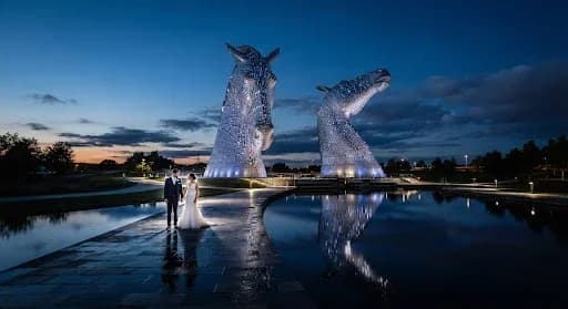 A wide-angle, breathtaking wedding photo taken at the Kelpies during dusk. The massive steel sculptures are illuminated with soft blue and white lights, while the sky is a deep indigo. The couple stands small in the frame, creating a sense of scale and modern majesty. The photography style is clean, sharp, and high-contrast, emphasizing the metallic textures and the romantic intimacy of the pair.