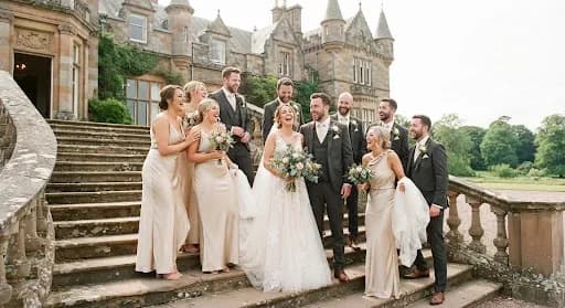 A candid, joyful moment of a wedding party laughing on the grand stone steps of Callendar House. The bridesmaids are in champagne-colored dresses that match the stone architecture perfectly. The sunlight is bright but soft, creating a high-key, airy feeling. The shot is natural and unposed, capturing authentic emotion in a high-fashion editorial style.