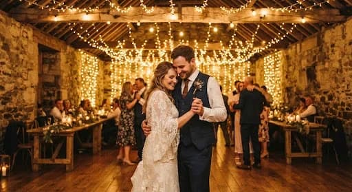 A romantic evening shot of a couple dancing under a canopy of fairy lights at a rustic Falkirk barn venue. The background is filled with warm, golden bokeh from the lights, while the couple is captured in a soft, ethereal glow. The image has a dreamlike quality, with rich shadows and sparkling highlights, emphasizing the magic of the celebration.