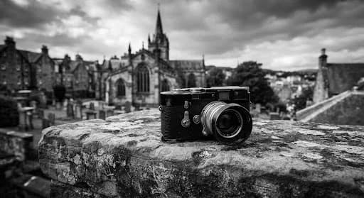 A detailed black and white portrait of a photographer's vintage Leica camera resting on a weathered stone ledge with the Church of the Holy Rude in the soft-focus background. The lighting is moody and dramatic, highlighting the textures of the stone and the polished metal of the camera. The image evokes a sense of history, craftsmanship, and a deep connection to Stirling's architectural heritage.