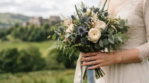 A candid, high-fashion close-up of a wedding bouquet featuring local Scottish thistles and cream roses, held by a bride whose hands are adorned with delicate heirloom jewelry. The background is a blurred tapestry of Stirling's lush green hills. The lighting is soft and natural, emphasizing the fine details and the romantic, high-end editorial style of the photography.