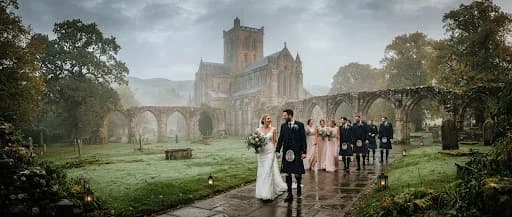 A breathtaking full-width landscape photograph of a wedding party walking through the misty grounds of Dunblane Cathedral near Stirling. The ancient stone arches are framed by ancient oak trees, and a soft morning fog clings to the grass. The composition is cinematic and expansive, capturing the ethereal beauty of the Scottish landscape and the elegance of the wedding attire in a high-contrast, fine-art style.