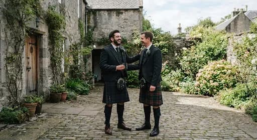 A dynamic, candid shot of a groom and his best man sharing a laugh in the courtyard of a historic Stirling townhouse. They are dressed in traditional Scottish kilts with charcoal jackets. The background shows the textured gray stone walls and a hint of a blooming garden. The lighting is bright and natural, capturing a genuine moment of joy and friendship in a high-end documentary style.