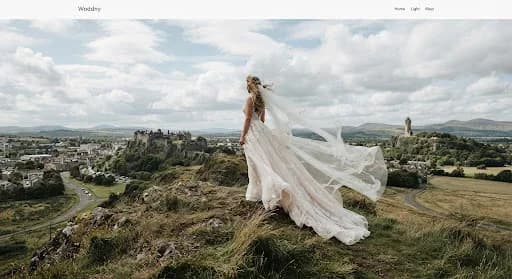 A dramatic shot of a bride's train flowing in the wind as she stands atop a grassy hill overlooking the Stirling skyline. The National Wallace Monument is visible in the distant haze. The sky is filled with voluminous white clouds against a pale blue backdrop. The image has a majestic, high-fashion quality, using the grand scale of the landscape to frame the delicate beauty of the wedding attire.